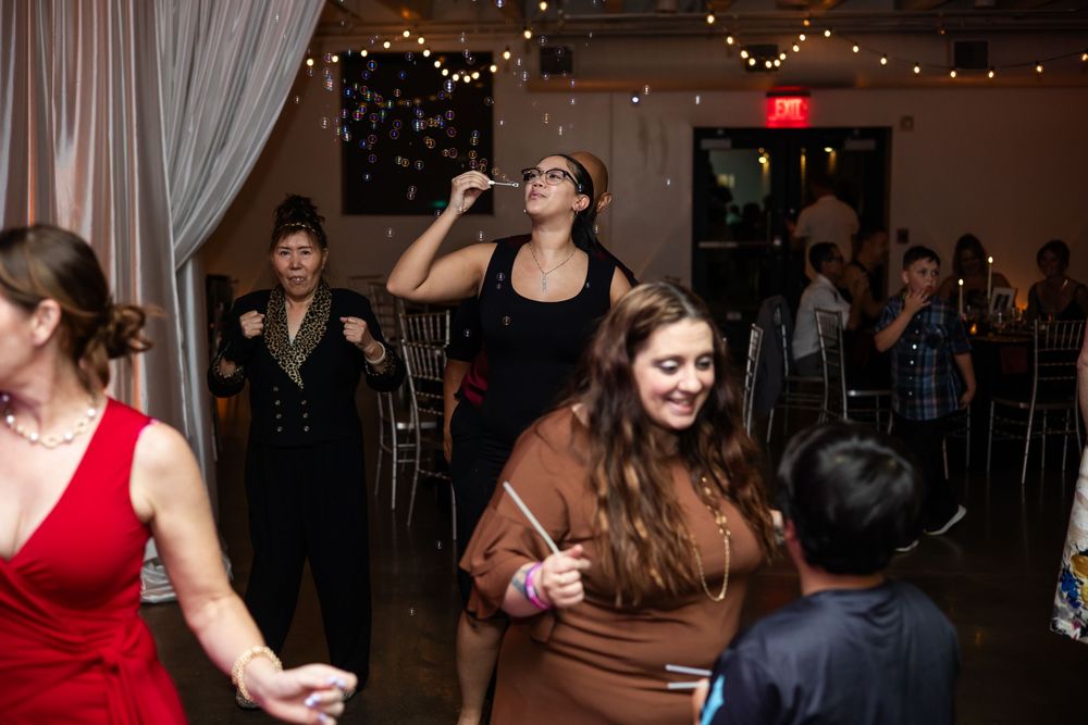 Guests laugh and blow bubbles on a dance floor at an indoor party with string lights and draped curtains.