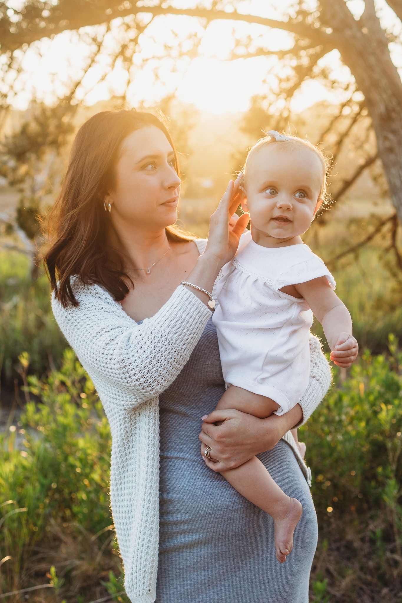 Mother in a white knit cardigan and gray dress holds her barefoot baby girl in a white outfit outdoors in warm golden sunset light.