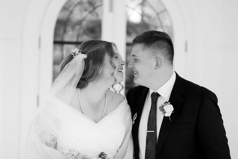 Black and white photo of a smiling bride in a veil and groom in a dark suit with boutonniere laughing together on their wedding day.