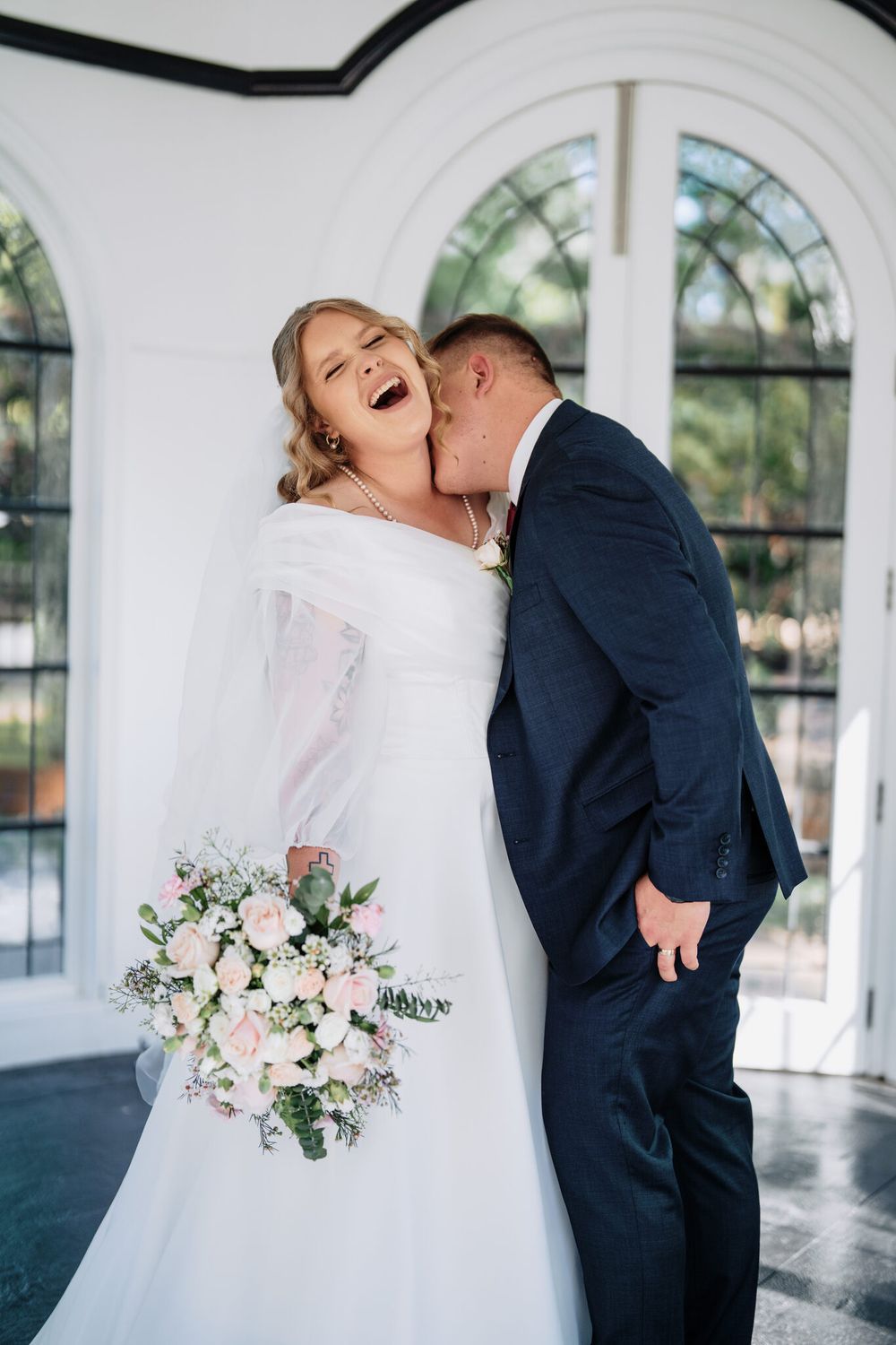 Laughing bride in a white off-shoulder wedding gown holds a pink rose bouquet as the groom in a navy suit kisses her cheek.