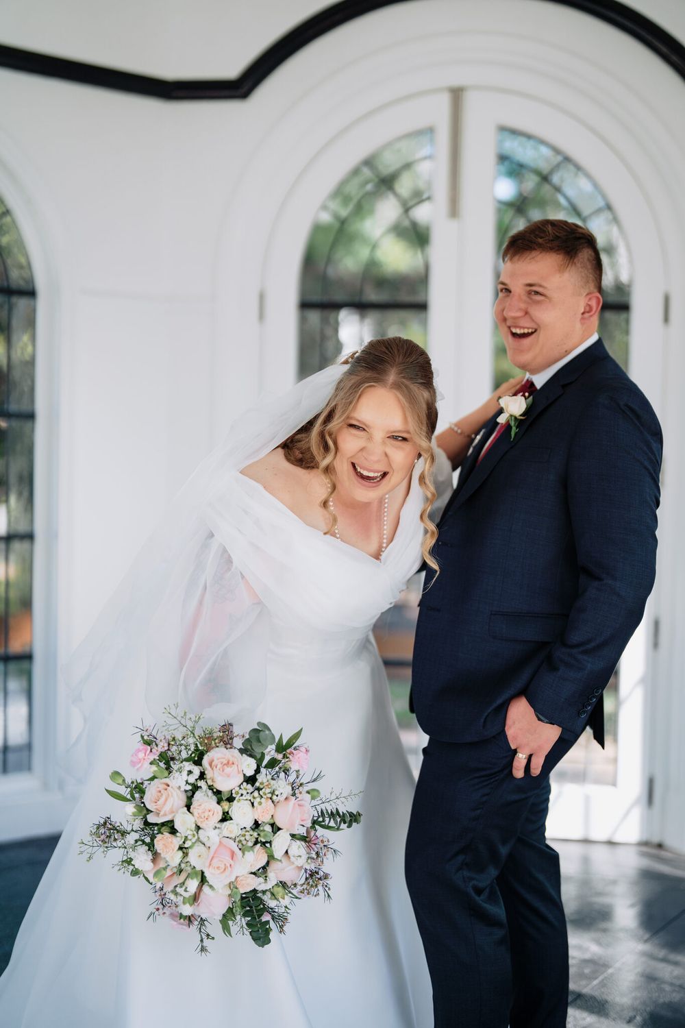 Laughing bride in an off-the-shoulder white gown holding a pink rose bouquet beside a smiling groom in a navy suit by arched windows.