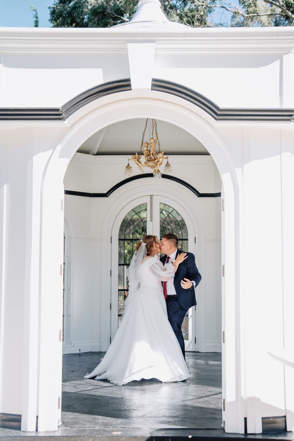 Bride in a white gown and groom in a navy suit share a kiss inside a white arched chapel with a gold chandelier.