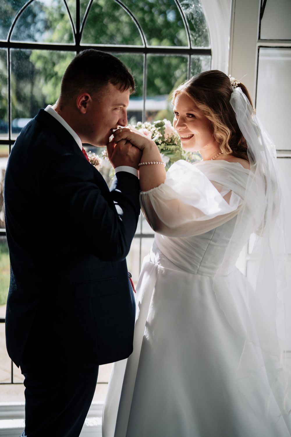 Groom in a dark suit kisses the hand of his smiling bride in a white gown and veil beside a large arched window.