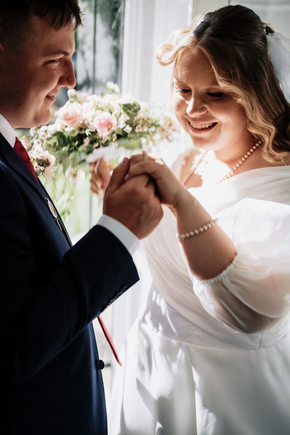 Smiling bride in a white dress and pearl necklace holds the groom's hand during a wedding ceremony, with a pink floral bouquet nearby.