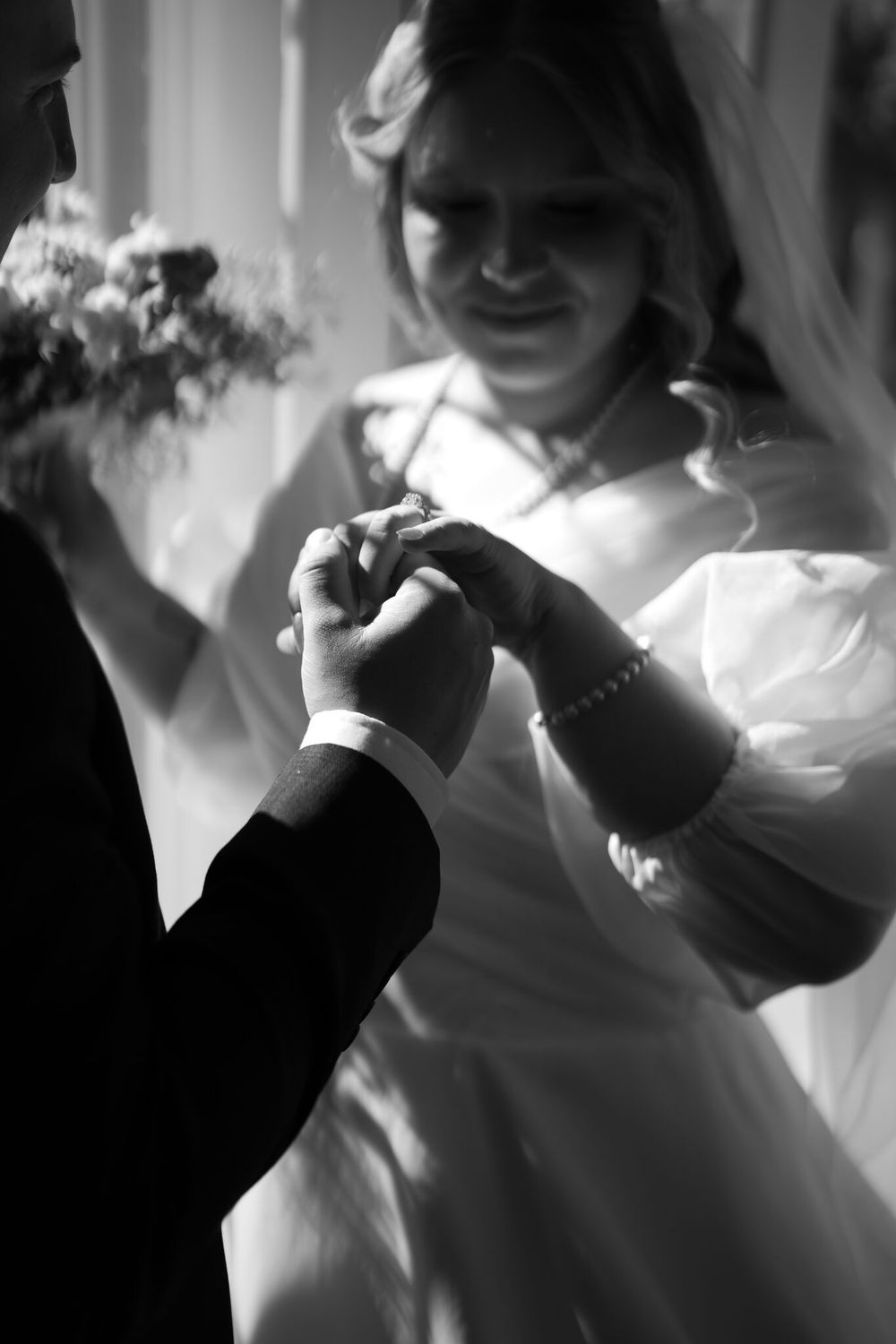 Black and white photo of a bride in a veil and puffed-sleeve gown exchanging wedding rings with the groom, holding a bouquet.