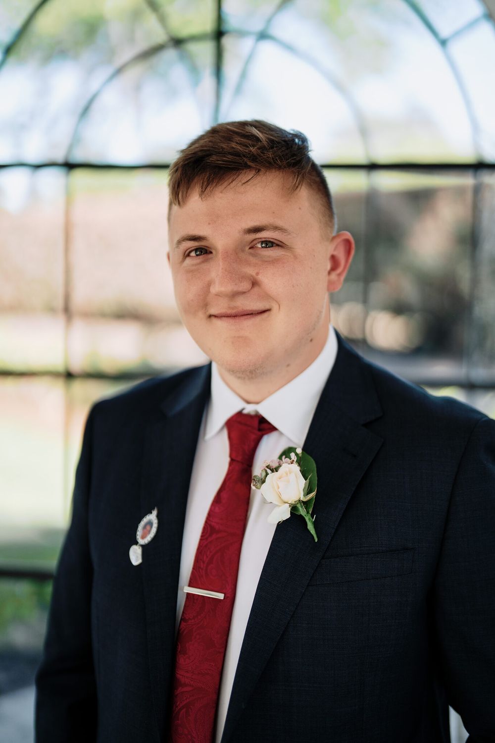 Smiling young man in a navy suit, white shirt, red patterned tie and white rose boutonniere standing in front of a glass-paneled window.