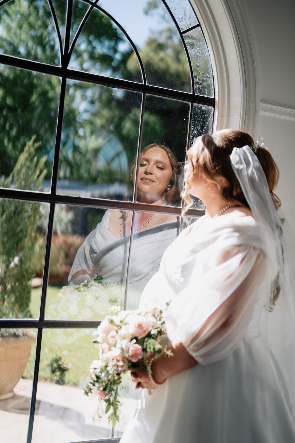 Bride in a white gown and veil holds a pink floral bouquet beside an arched window, her reflection glowing in the glass.
