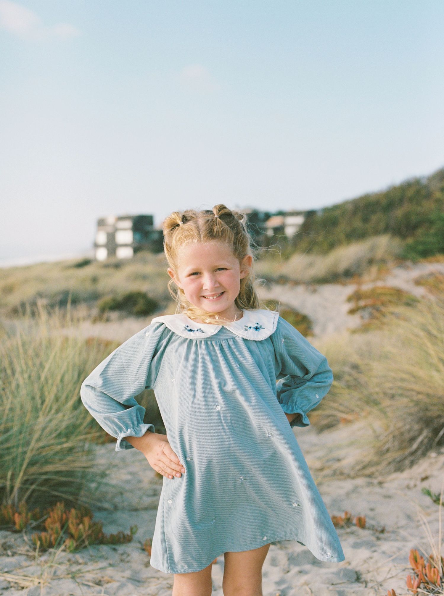 Smiling young girl in a blue embroidered collar dress stands with hand on hip among beach grass and sand dunes at sunset.