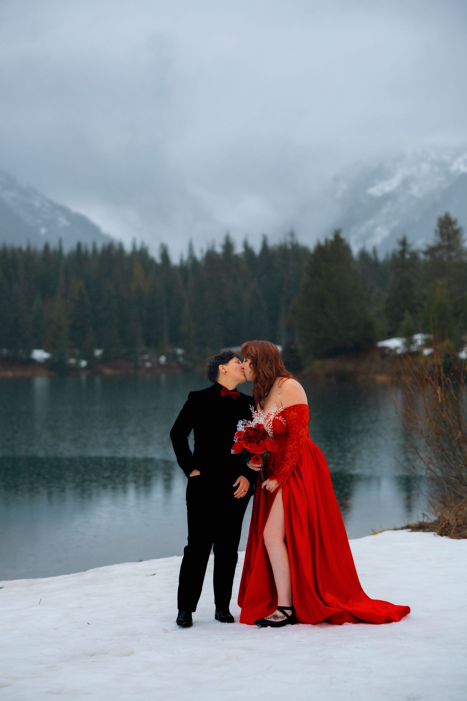 Groom in black suit lifts smiling bride in white dress and veil in front of a scenic waterfall during outdoor wedding photos.
