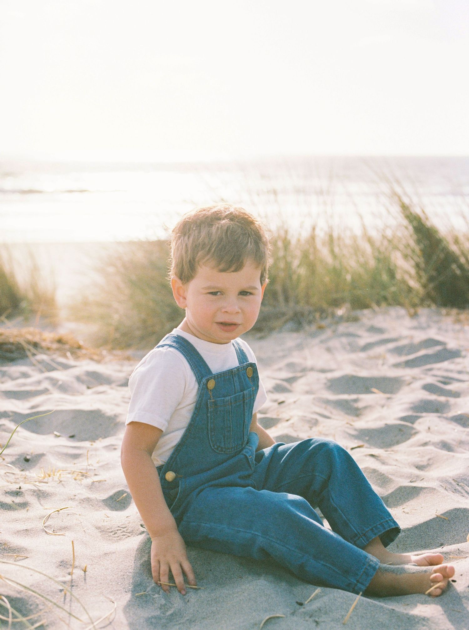 Toddler in denim overalls and a white tee sits barefoot on a sandy beach with soft dune grass and the ocean glowing in golden light.