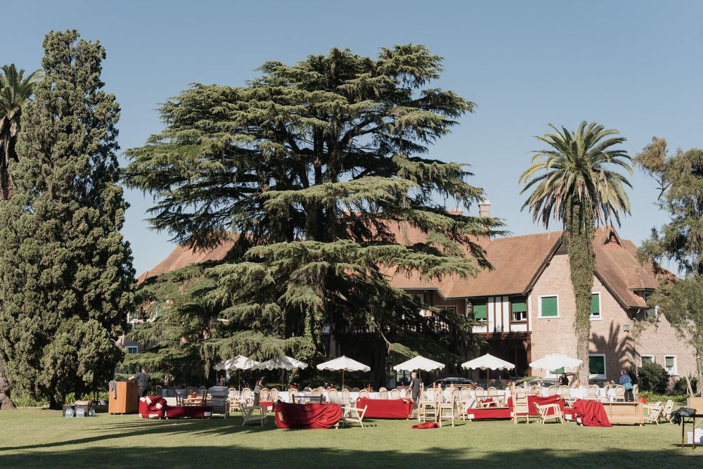 Garden wedding setup with red-draped tables and umbrellas on a lawn before a stately house and a large cedar tree.