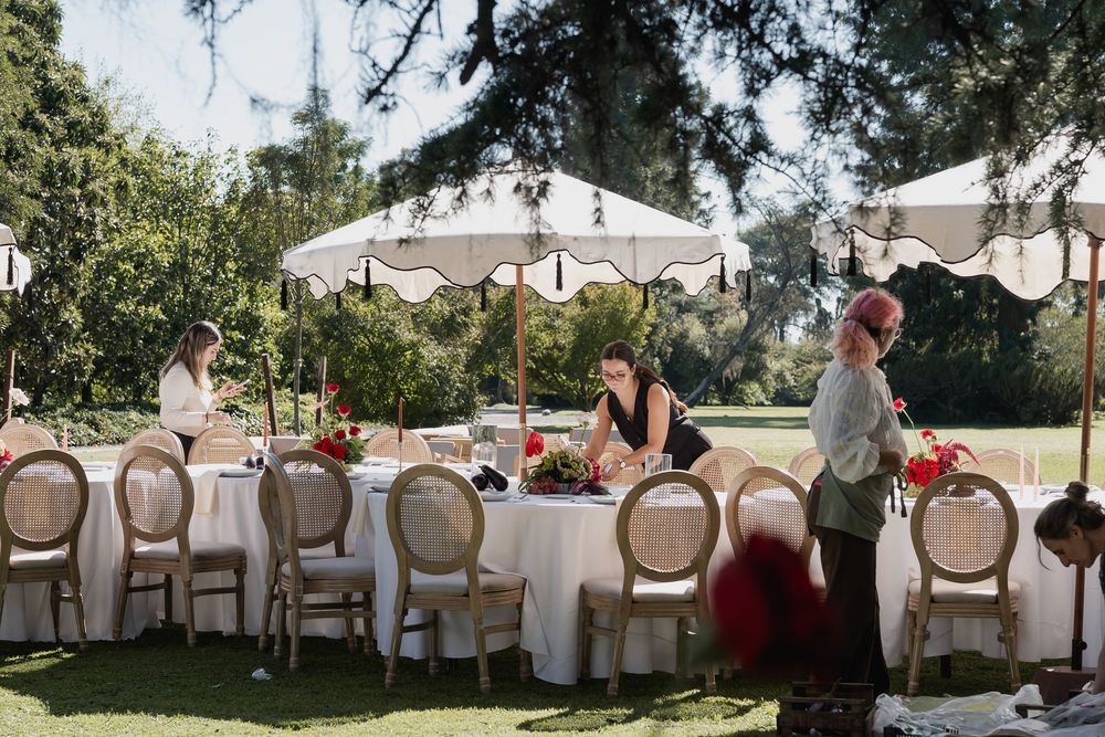 Wedding staff arrange floral centerpieces on a long table set with rattan chairs and white fringed umbrellas in a leafy garden.