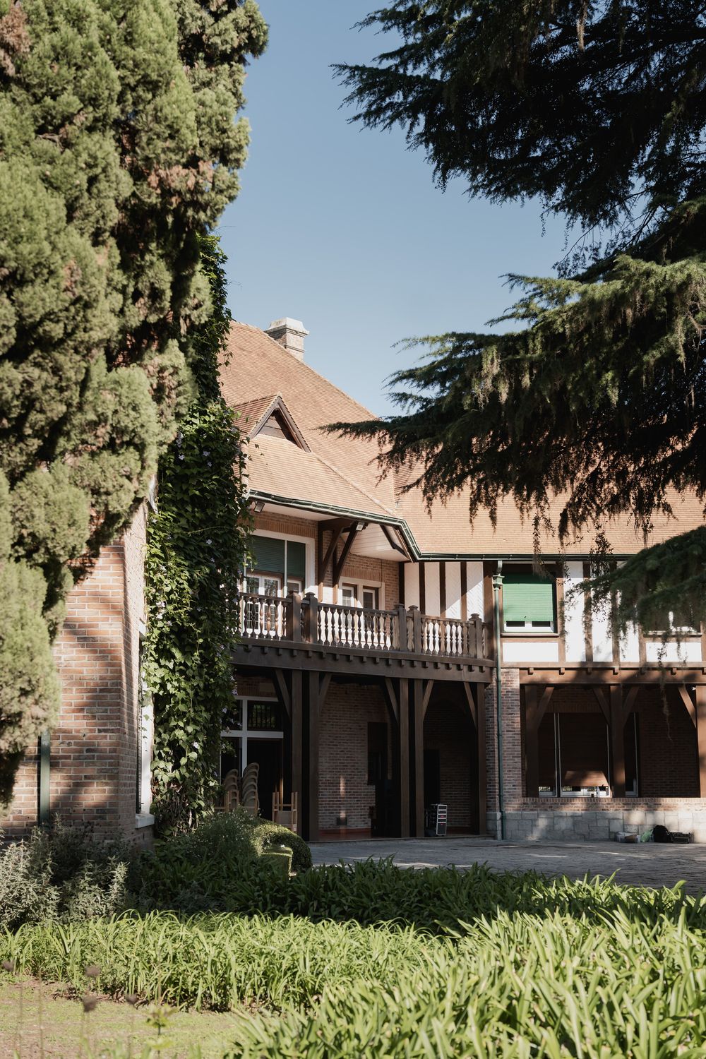 Tudor-style brick house with timber framing, a wooden balcony and terracotta roof, surrounded by tall cypress trees and a lush garden.