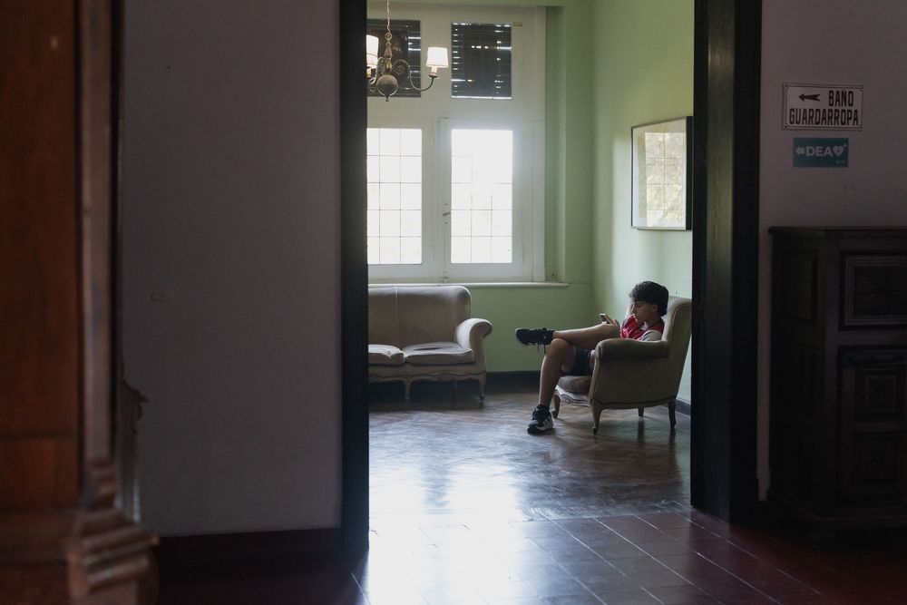 Person sits on an armchair in a quiet, sunlit room with green walls and wood floors, viewed through a dark doorway.