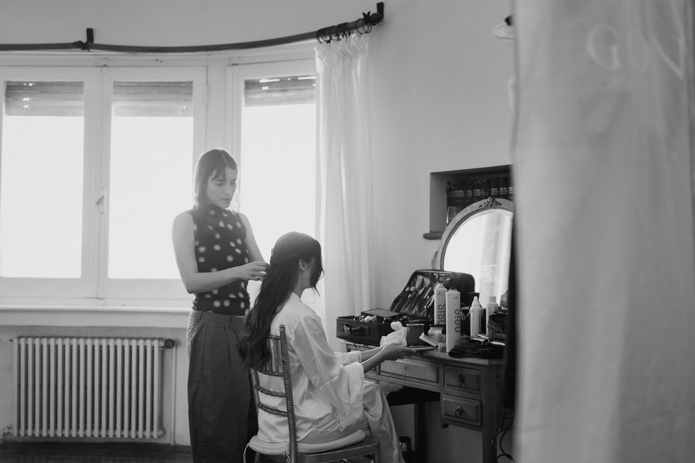 A bridesmaid stands beside a seated bride at a vanity as she prepares her hair and makeup in a bright room before the wedding.