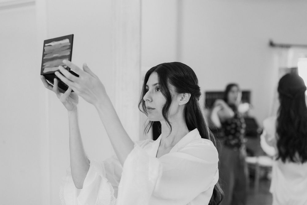 Black and white image of a bride holding a small mirror to check her hair and makeup while getting ready.
