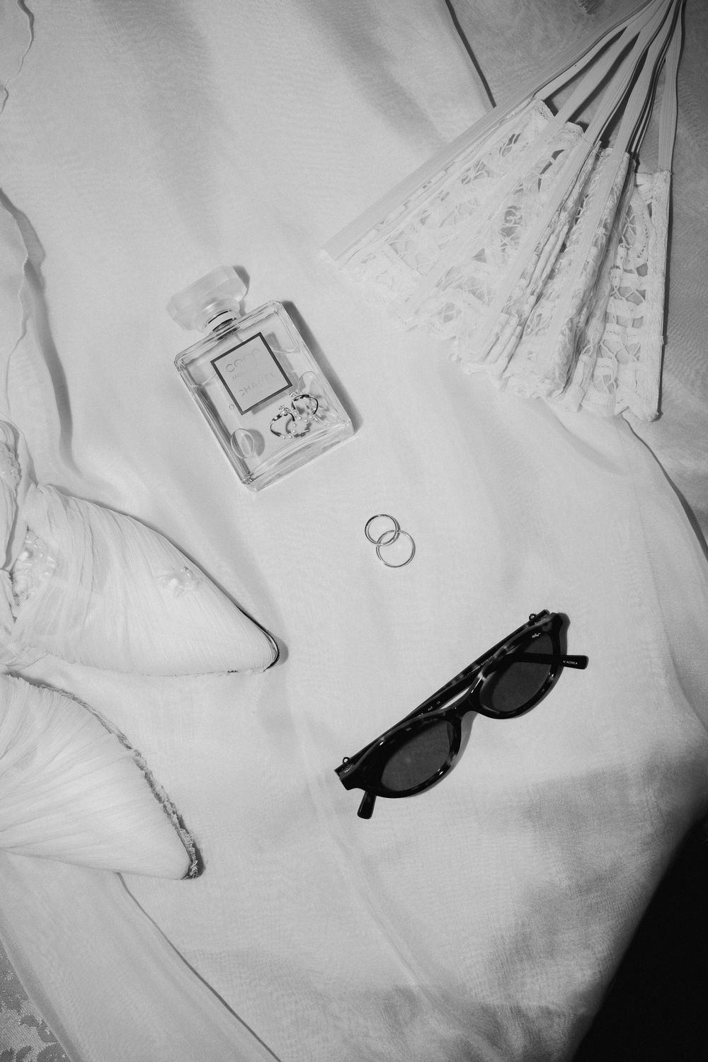 Black and white flat lay of wedding rings, perfume bottle, cat-eye sunglasses, lace fan and bridal shoes on white fabric.