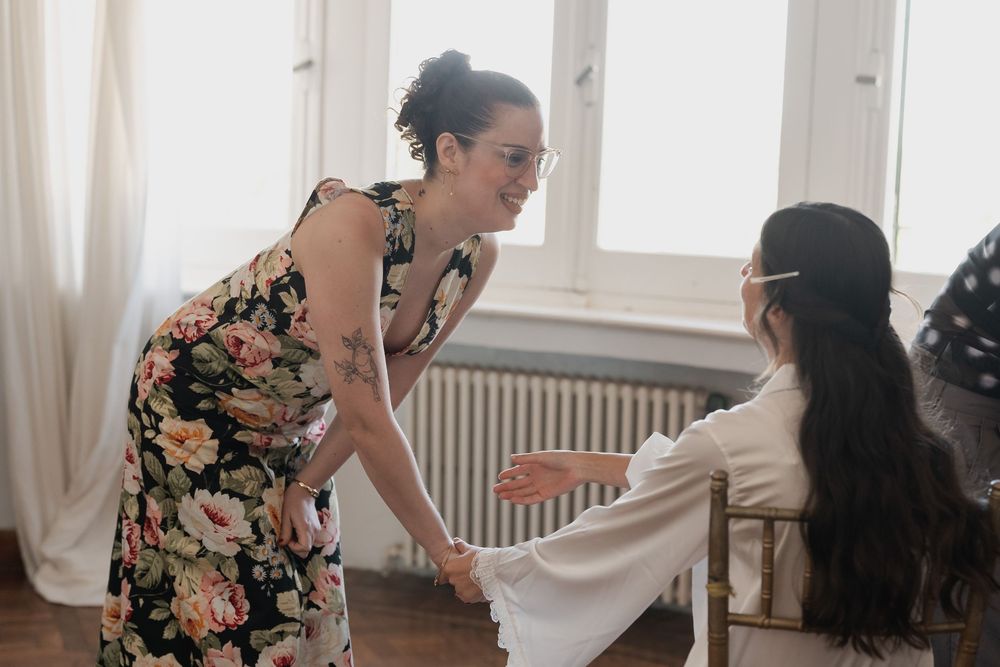 A woman in a floral dress smiles and holds hands with a seated bride in a white robe in a bright room before the wedding.