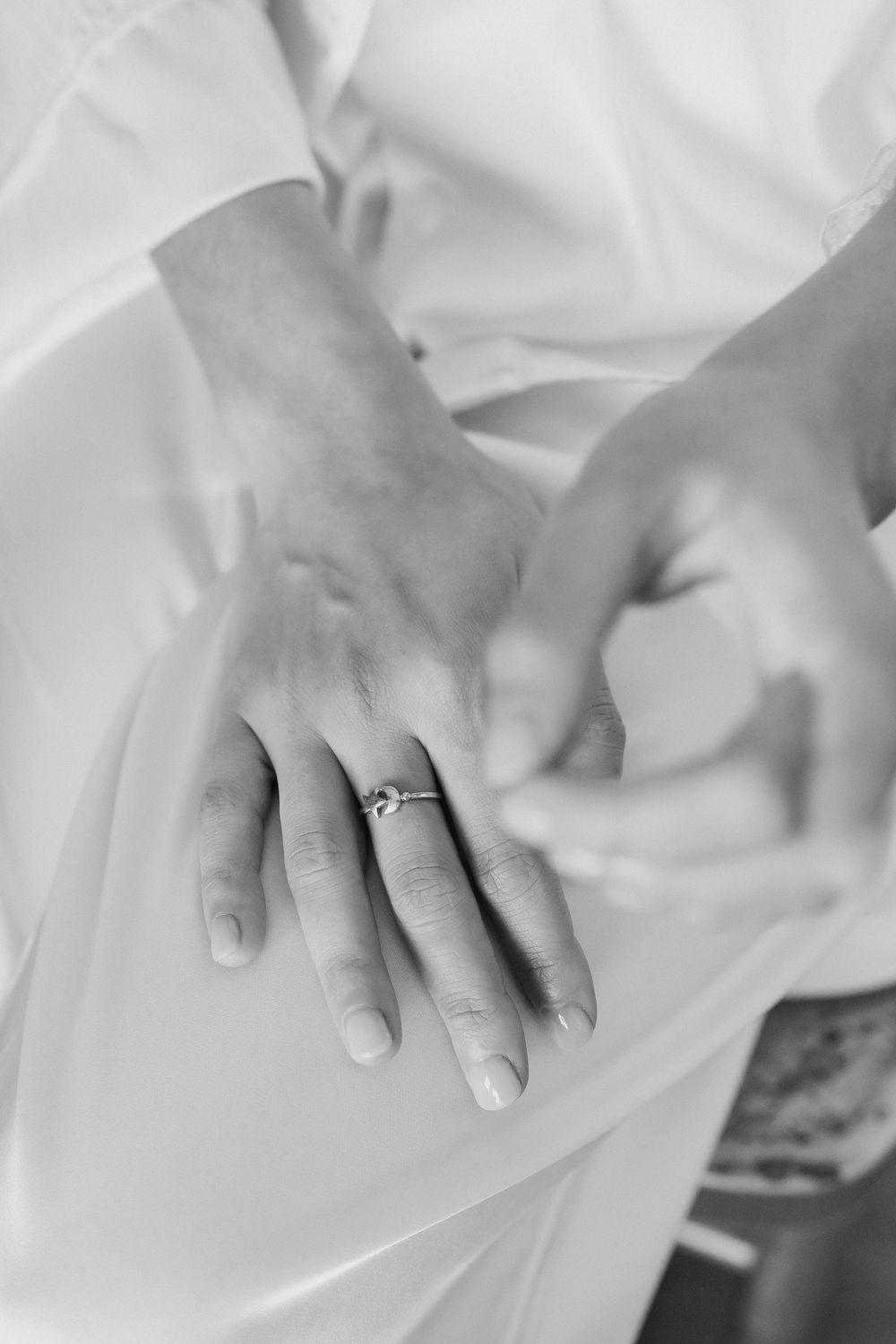 Black and white close-up of a bride's hand wearing a delicate diamond engagement ring, resting on her flowing silk robe.