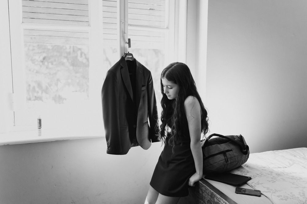 A young woman in a black dress leans against a window ledge near a hanging suit jacket and bag in soft natural light.