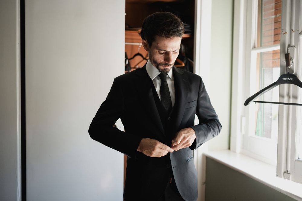 Groom in a dark suit and tie buttons his jacket while getting ready beside a bright window.