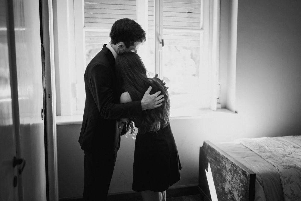 Black and white photo of a couple embracing beside a sunlit window in a quiet room with a bed in the corner.