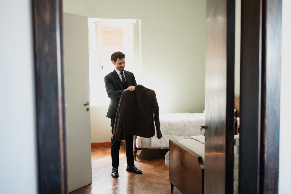 Groom in a dark suit and tie holds his jacket while standing in a sunlit bedroom as he gets ready for the wedding.