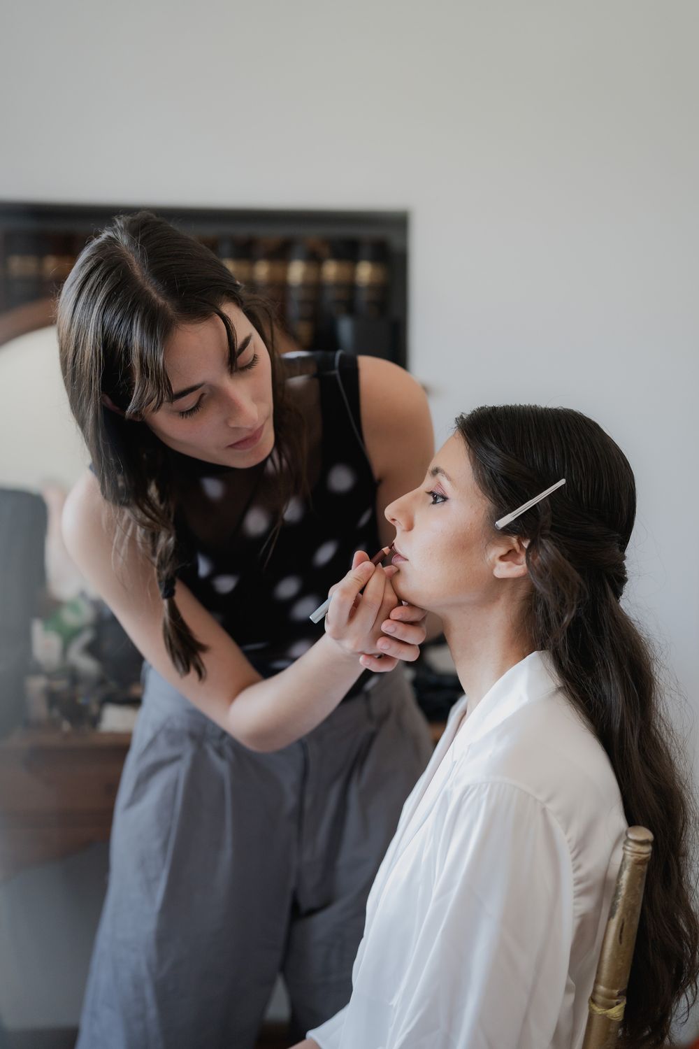 Makeup artist applies lipstick to a bride in a white robe during bridal preparations in a bright room.