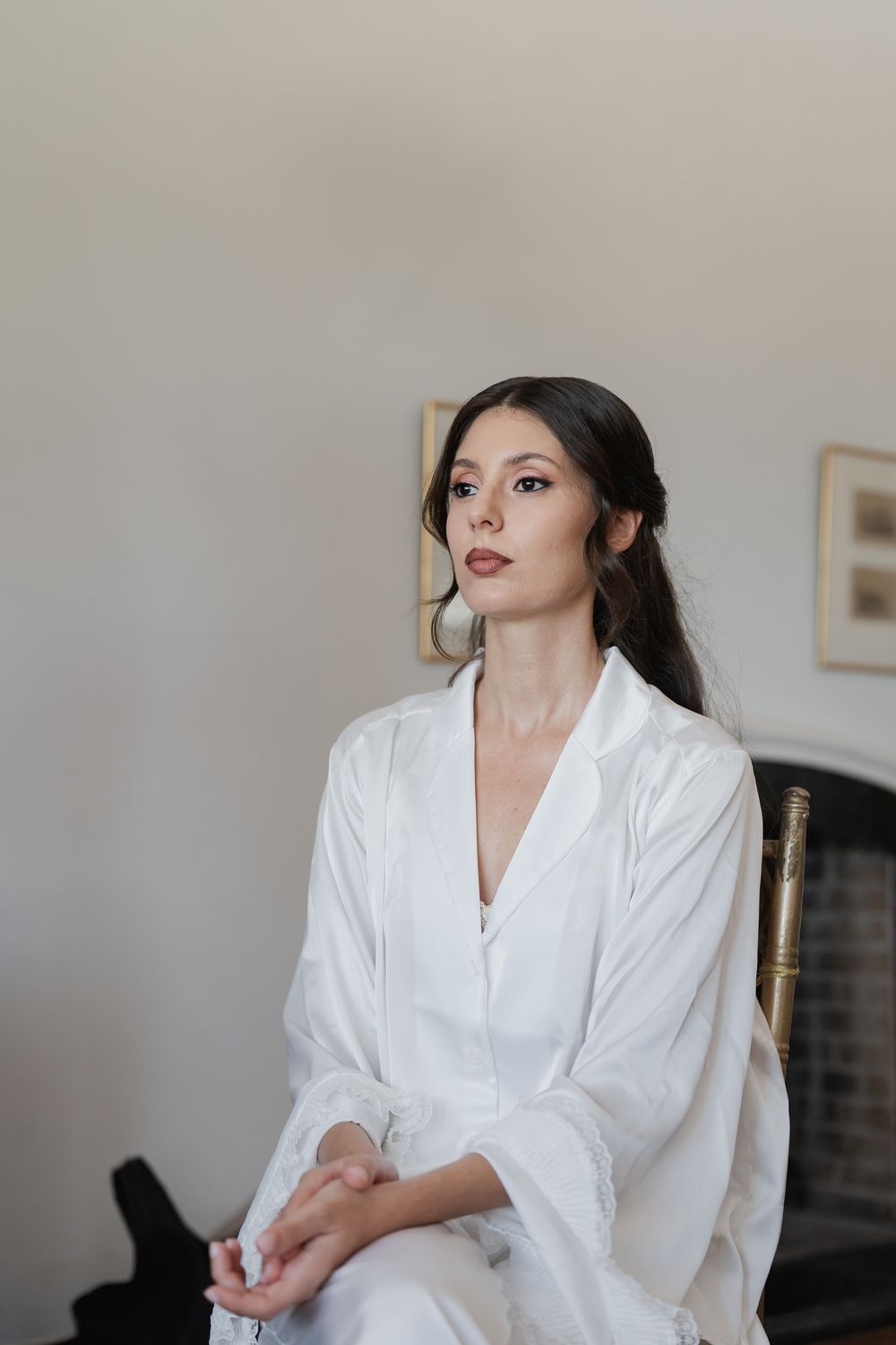 A bride in a white satin robe sits calmly on a gold chair with her hands folded, her hair styled in an elegant updo.