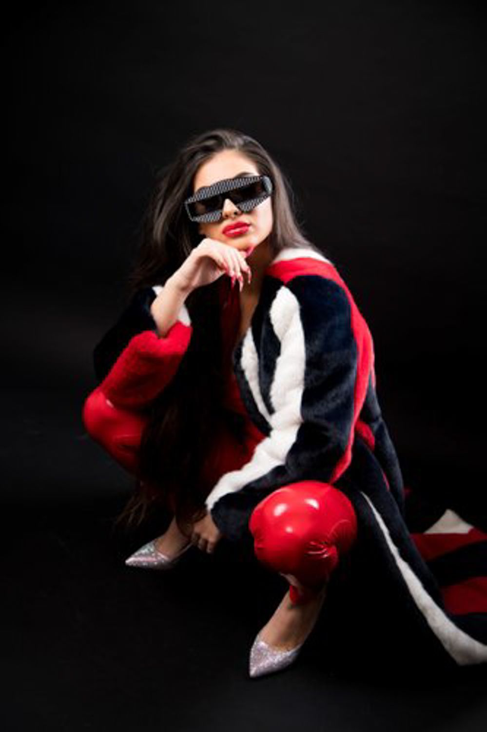 Woman crouching in a red, white and black color-block faux fur coat with red boxing gloves, black sunglasses and glitter heels.