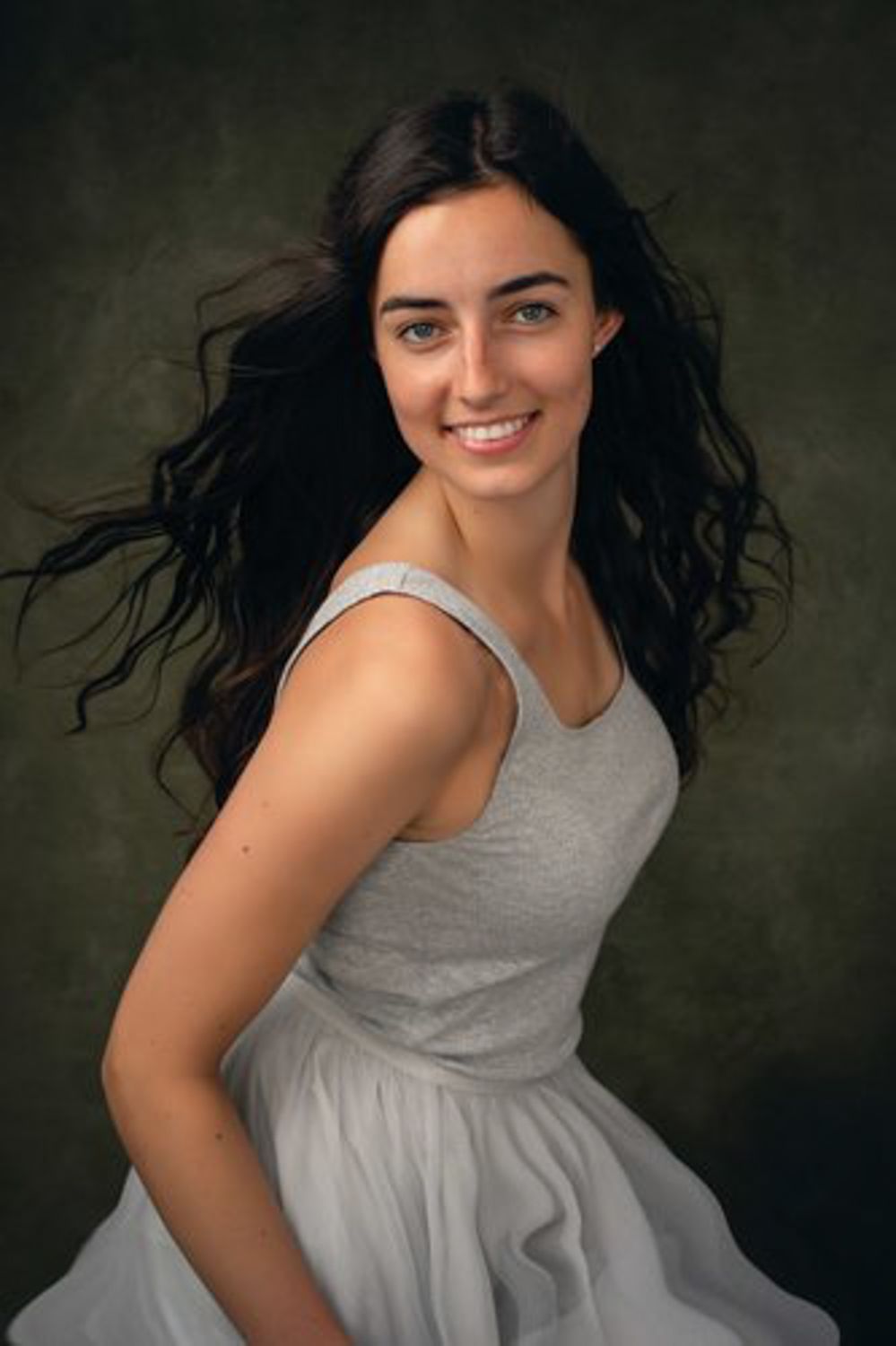 Smiling young woman with long dark wavy hair in a gray sleeveless tulle dress posing against a textured dark green studio backdrop.
