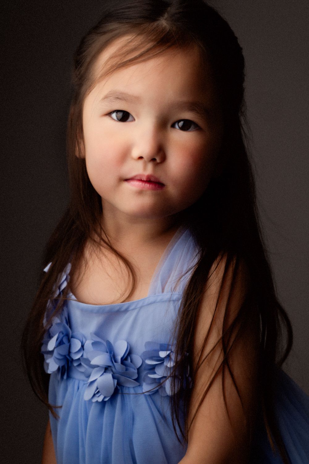 Studio portrait of a young girl with long dark hair wearing a blue tulle dress with floral appliqué against a dark background.