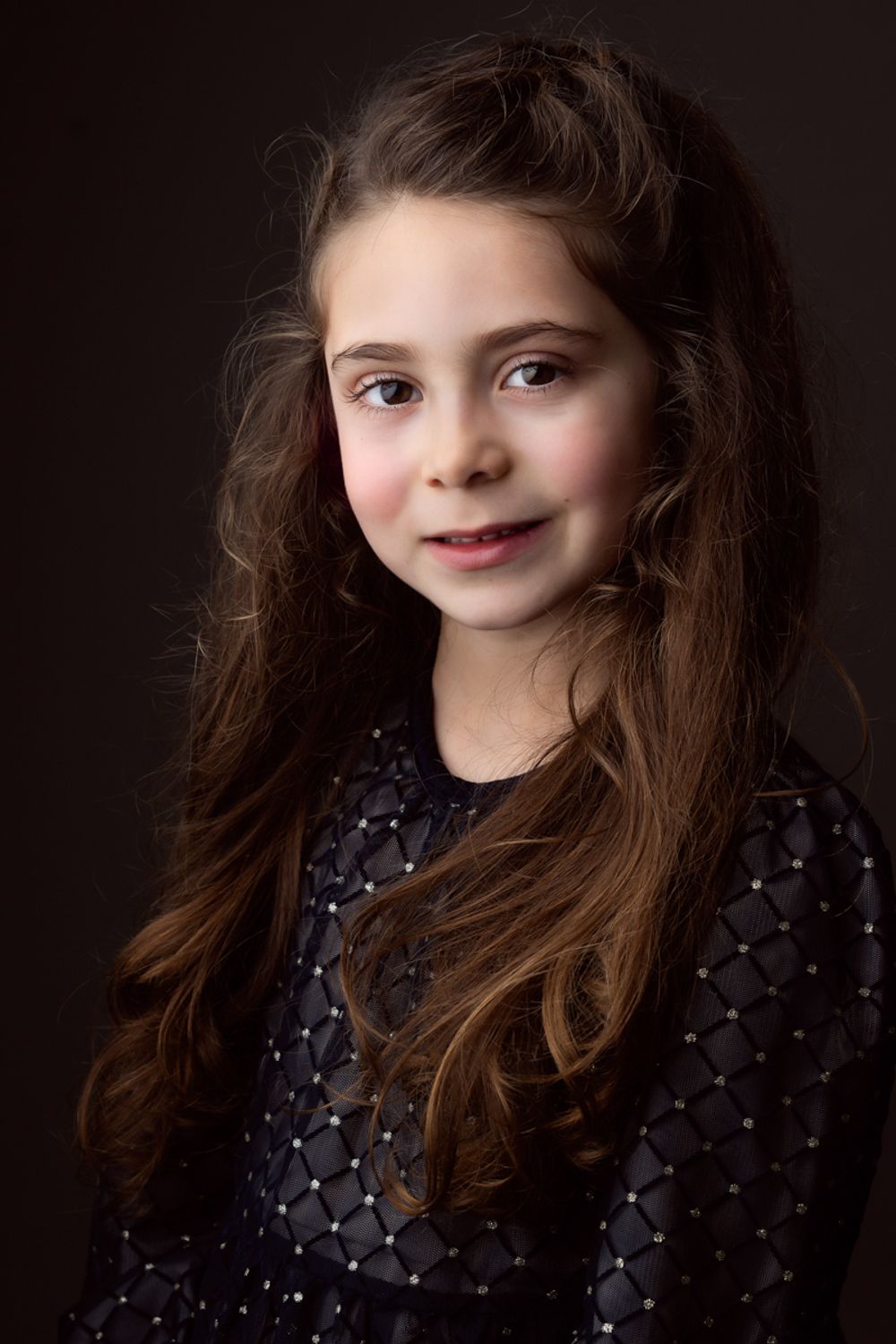 Studio portrait of a young girl with long wavy brown hair, wearing a dark sparkly diamond-patterned dress against a dark background.