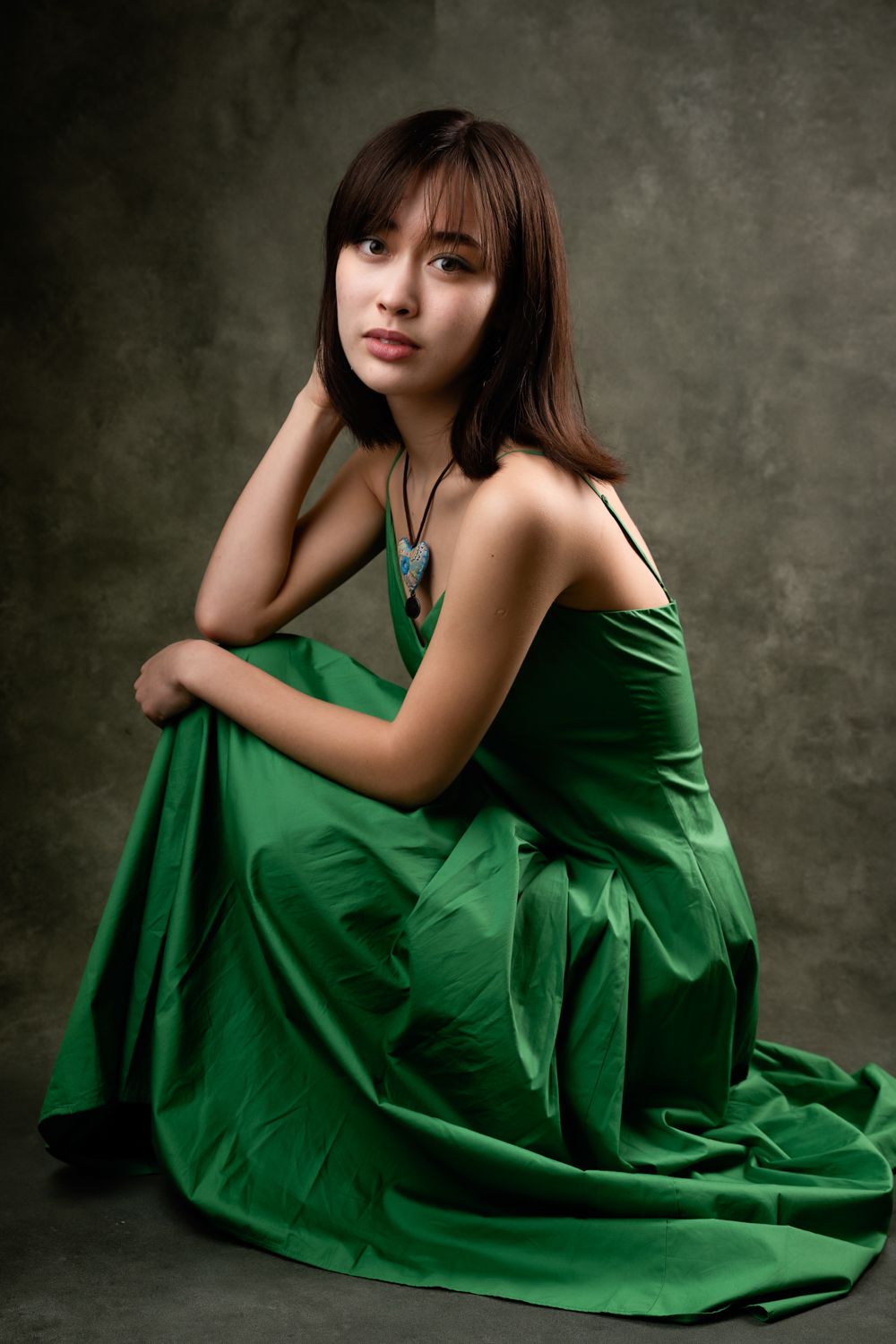 Young woman with shoulder-length brown hair and a pendant necklace poses seated in a flowing green dress against a textured gray backdrop.