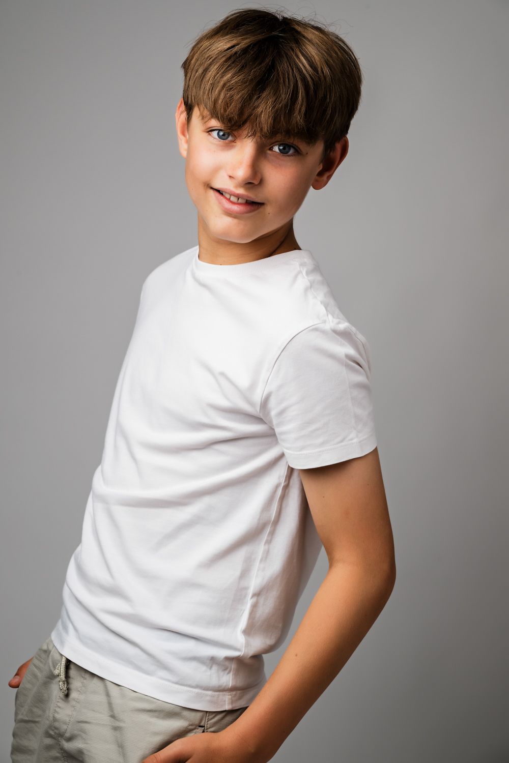 Smiling boy with short brown hair poses sideways in a white t-shirt and khaki shorts against a plain gray studio background.