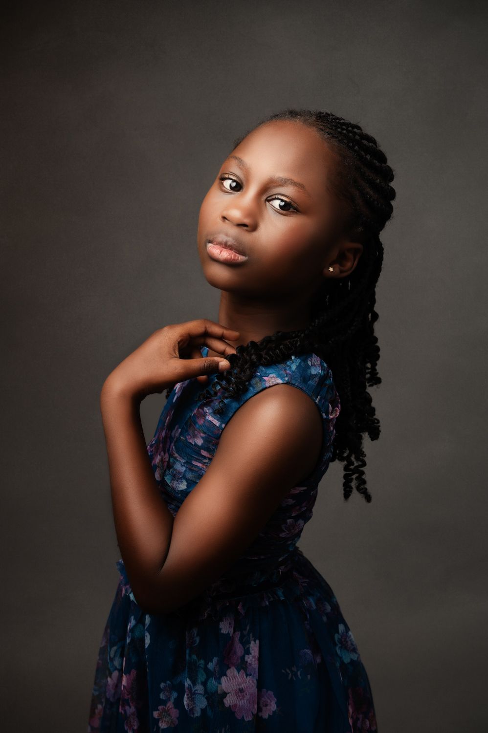 Young girl with long braided hair strikes a graceful studio pose in a blue floral sleeveless dress against a dark backdrop.