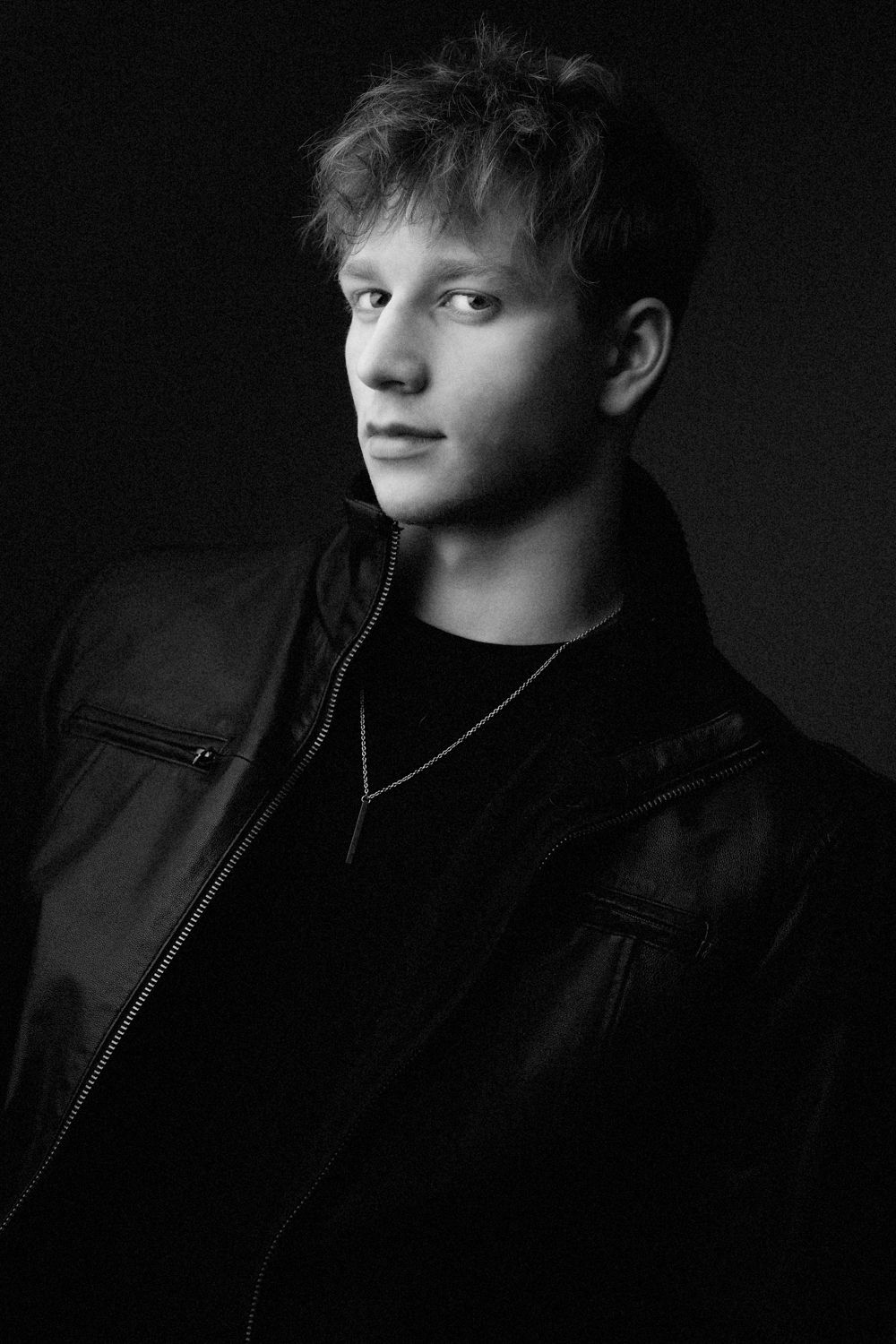 Black and white studio portrait of a young man with tousled hair in a leather jacket and pendant necklace against a dark background.