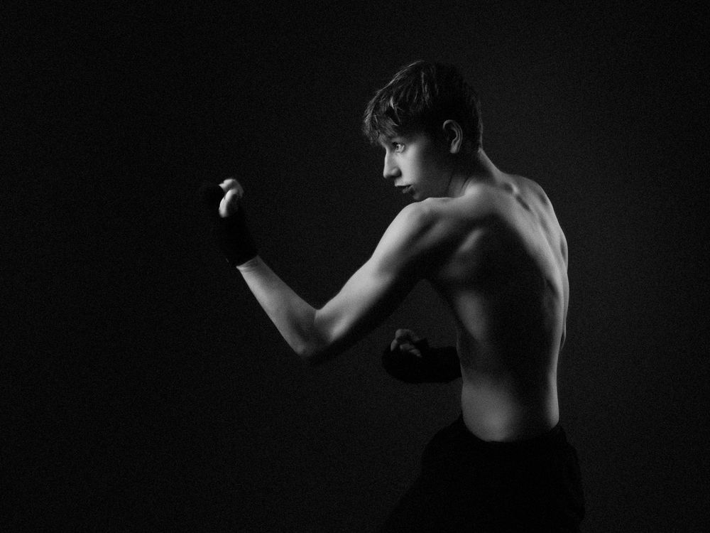 Black and white portrait of a shirtless young boxer in a fighting stance with wrapped hands against a dark studio background.