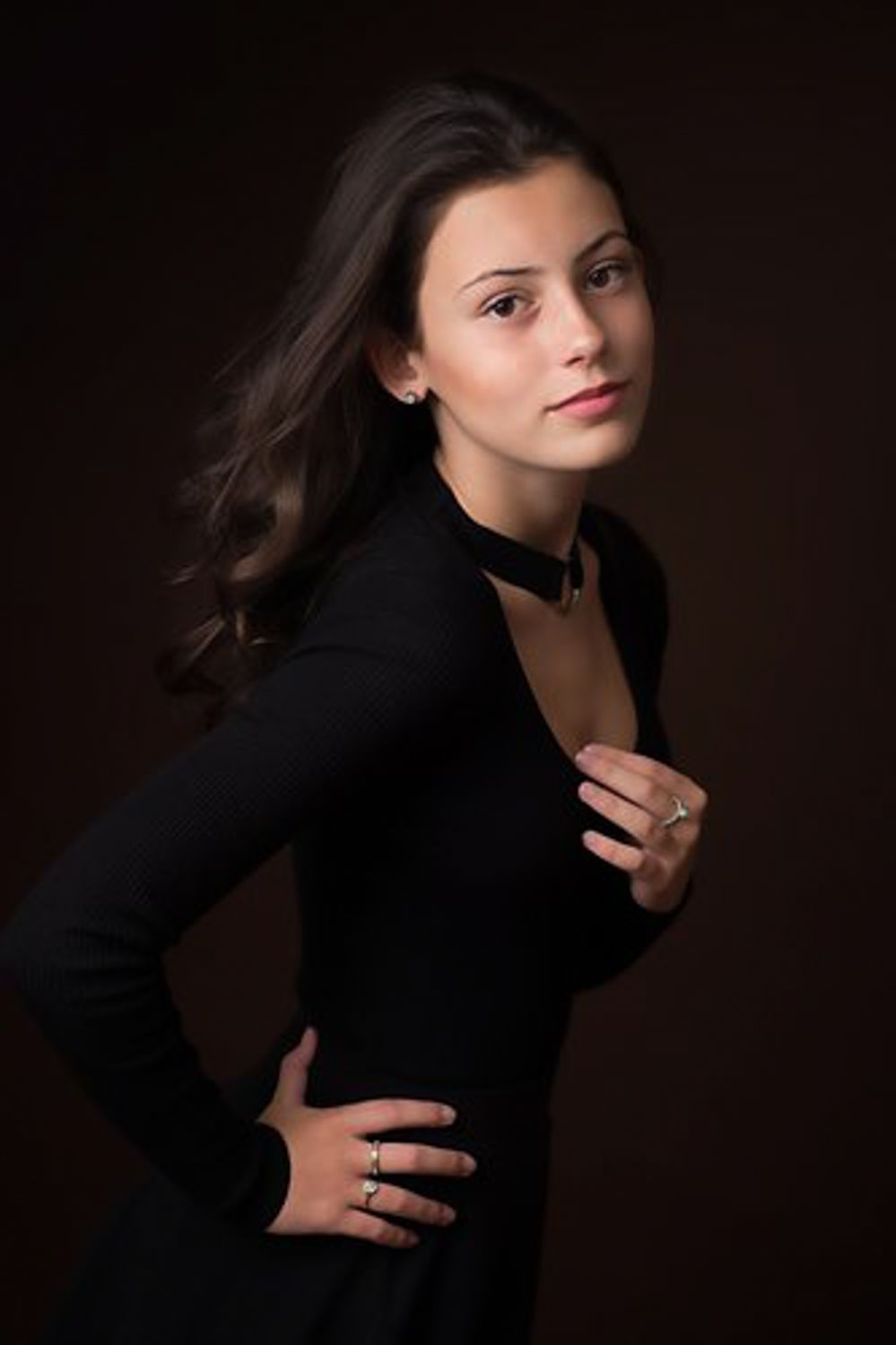 Studio portrait of a young woman with long dark hair in a black long-sleeve dress and choker, posing with a hand on her hip.