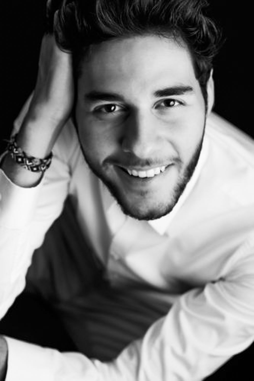 Black and white portrait of a smiling young man with short dark hair and a beard, wearing a white shirt and beaded bracelet.