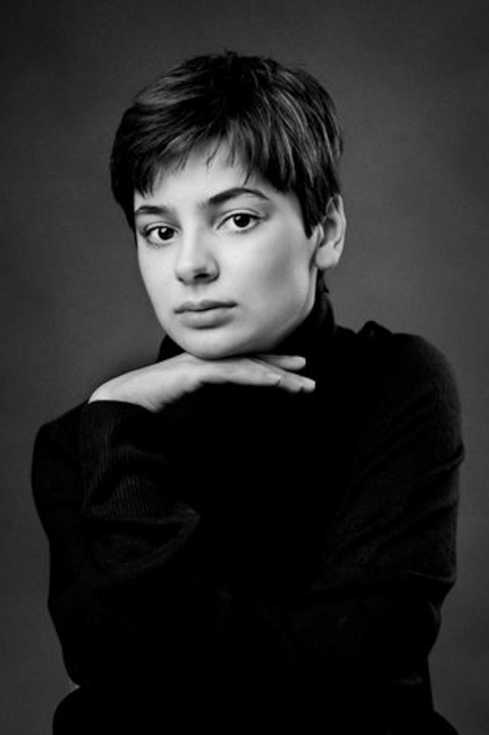Black and white studio portrait of a young woman with short dark hair resting her chin on her hand, wearing a dark turtleneck sweater.