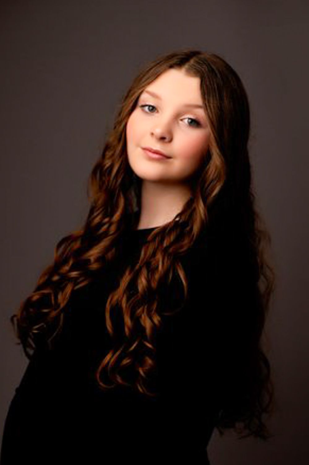 Studio portrait of a teenage girl with long curly auburn hair, wearing black and tilting her head against a muted gray backdrop.