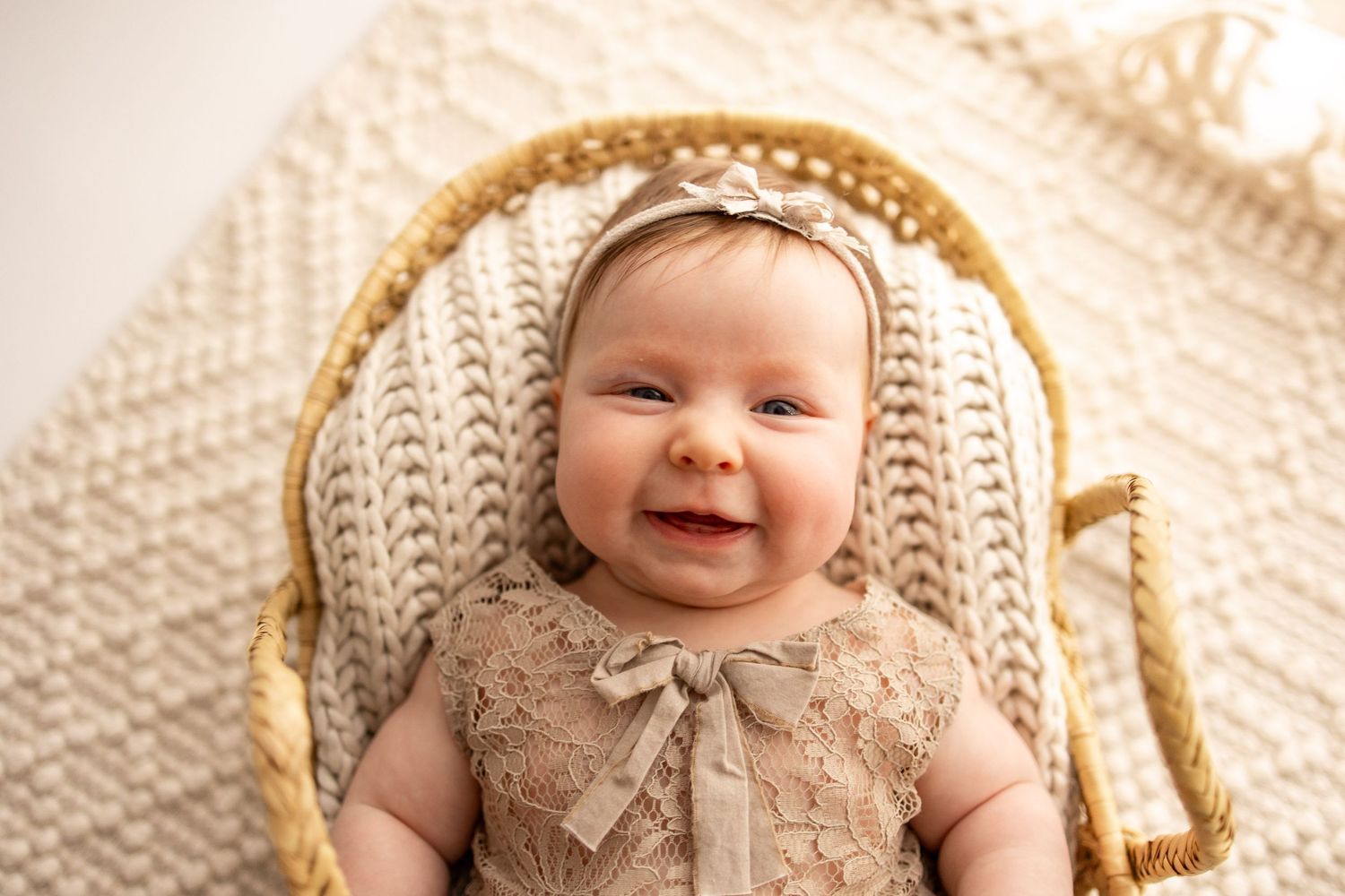 Smiling baby girl in lace romper and bow headband lying in a wicker Moses basket on a knit blanket.