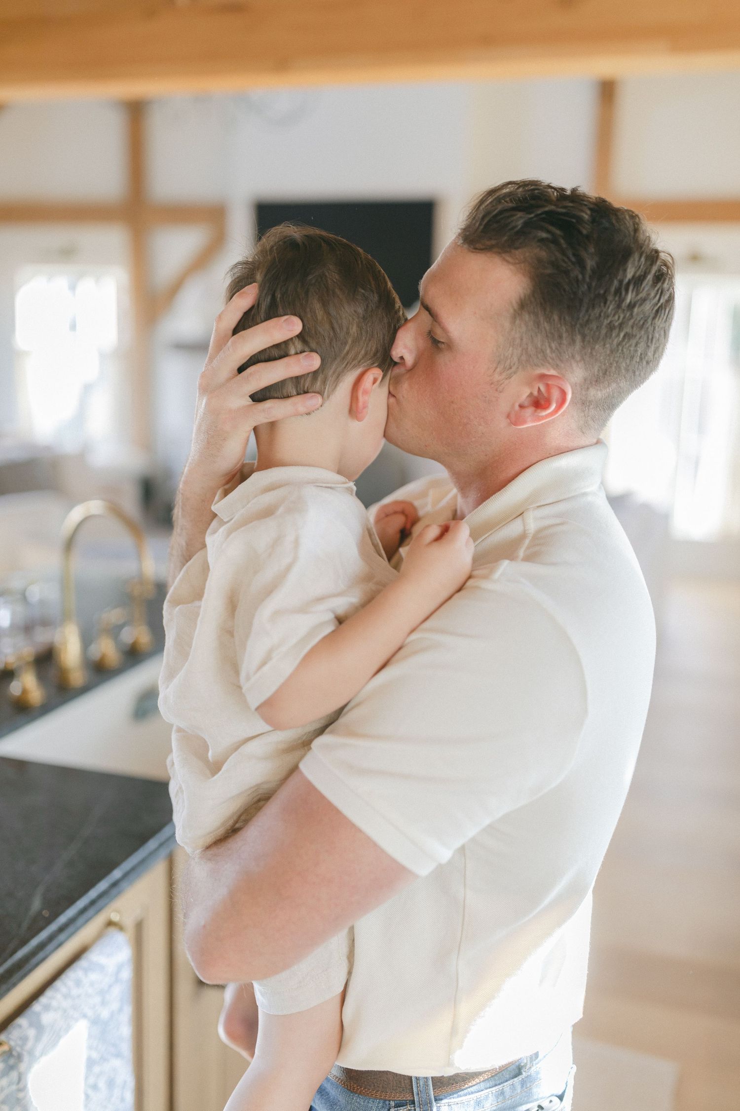 A father tenderly kisses his young child while holding him in a bright, modern kitchen.