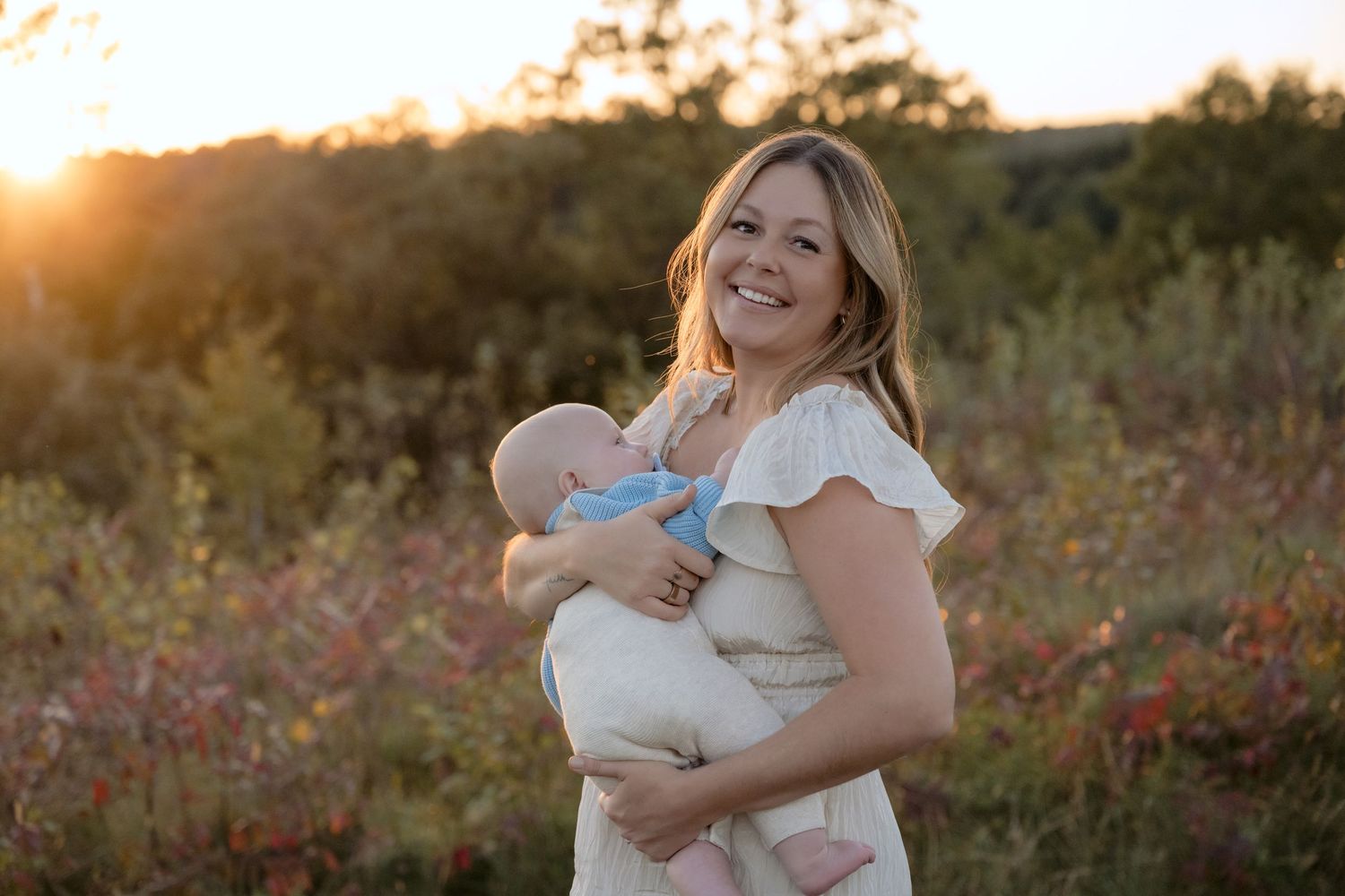 a young mom smiles with infant in her arms as the sunset glows behind her during fall mini session in hudson wisconsin