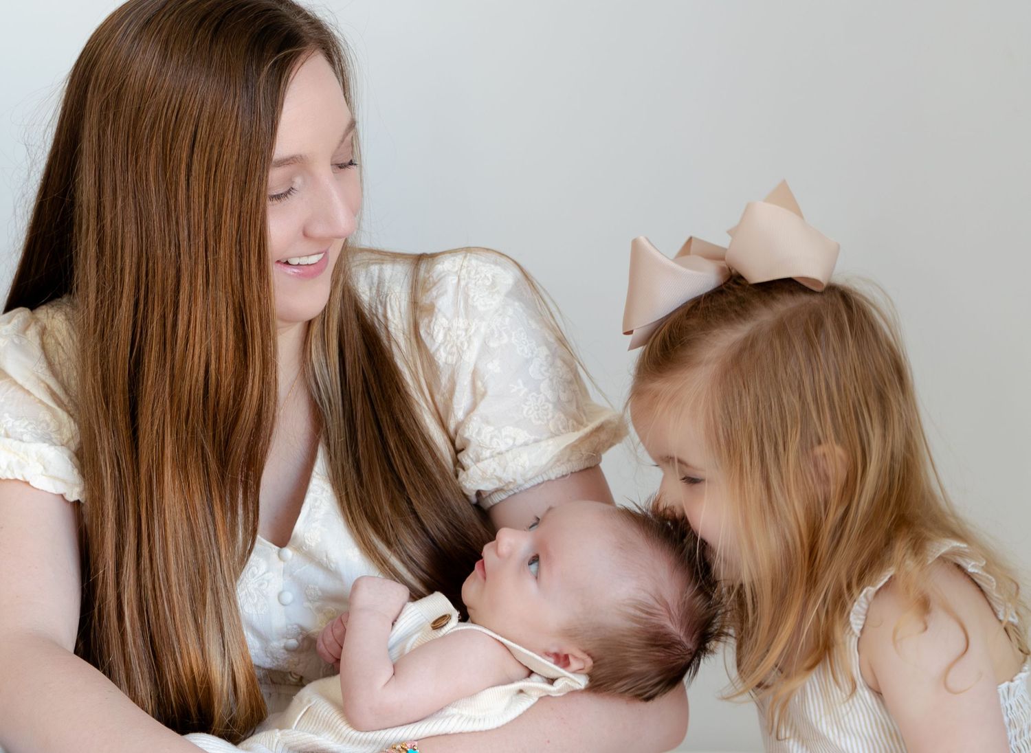 A mother smiles at her newborn baby while a young girl in a bow gives the infant a gentle kiss.