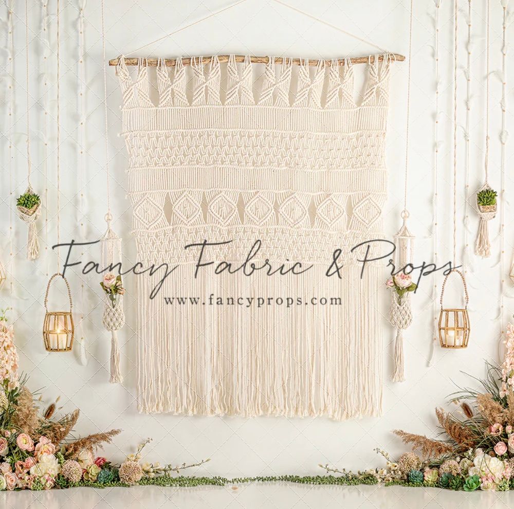 Boho macrame wall hanging backdrop with fringe, lanterns, and floral arrangements by Fancy Fabric & Props.