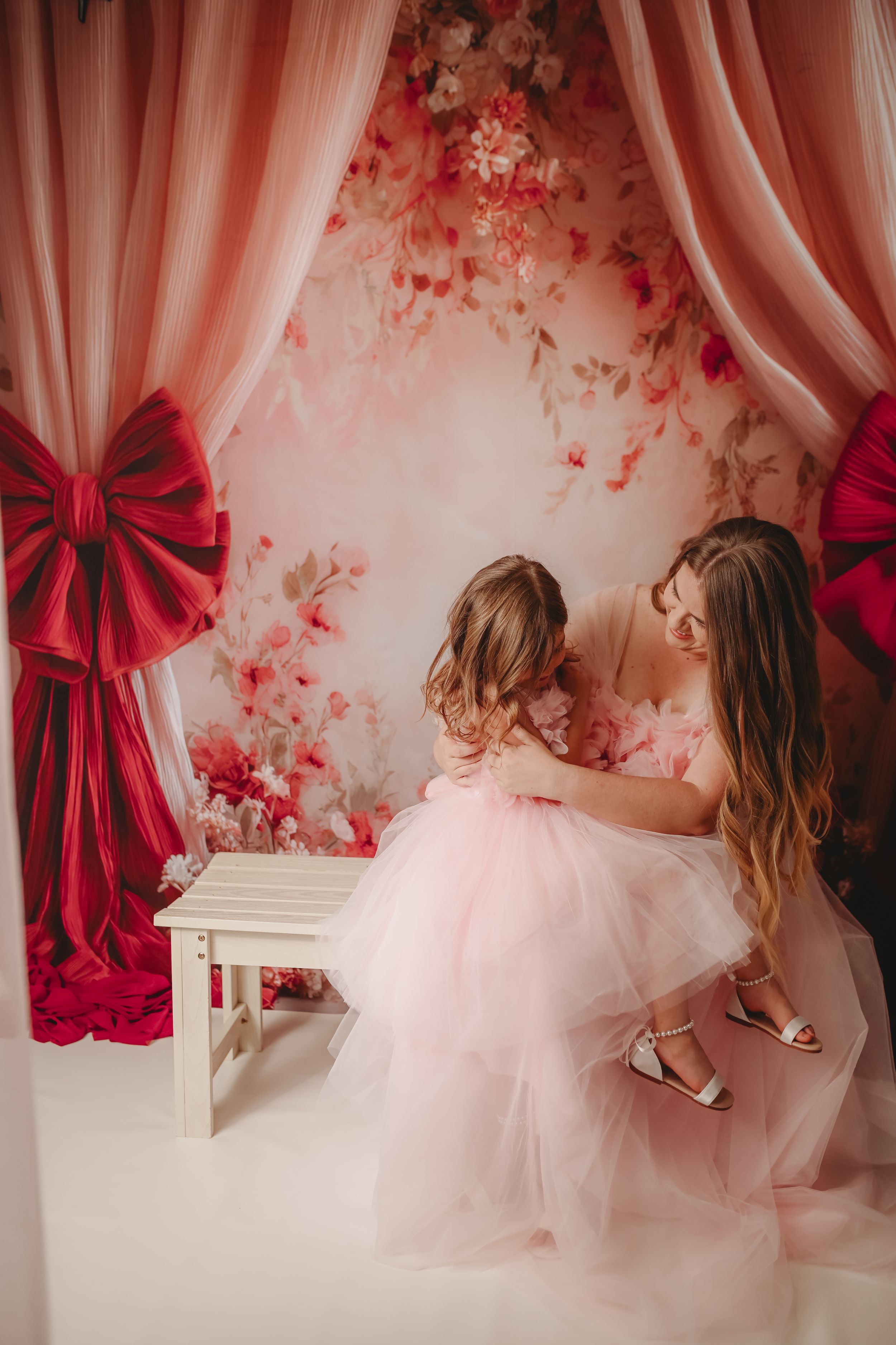 Mother and daughter in pink tulle dresses pose together in a floral backdrop studio with red bow curtains.