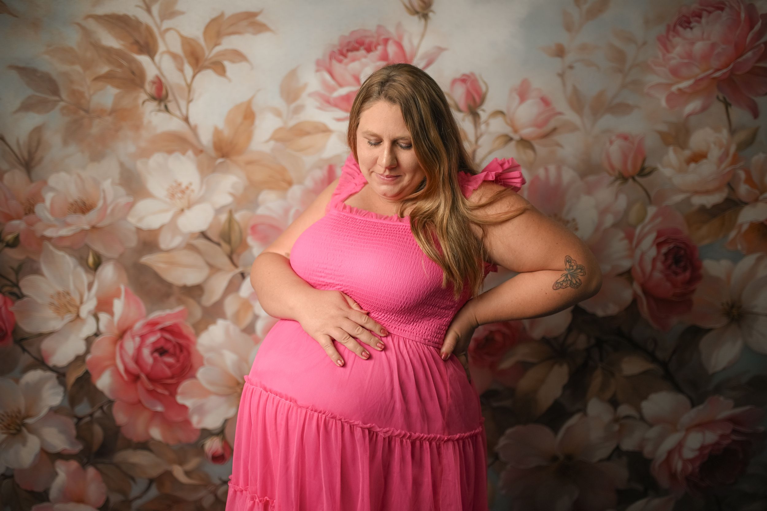 Pregnant woman in pink dress posing against floral backdrop during maternity photo session.