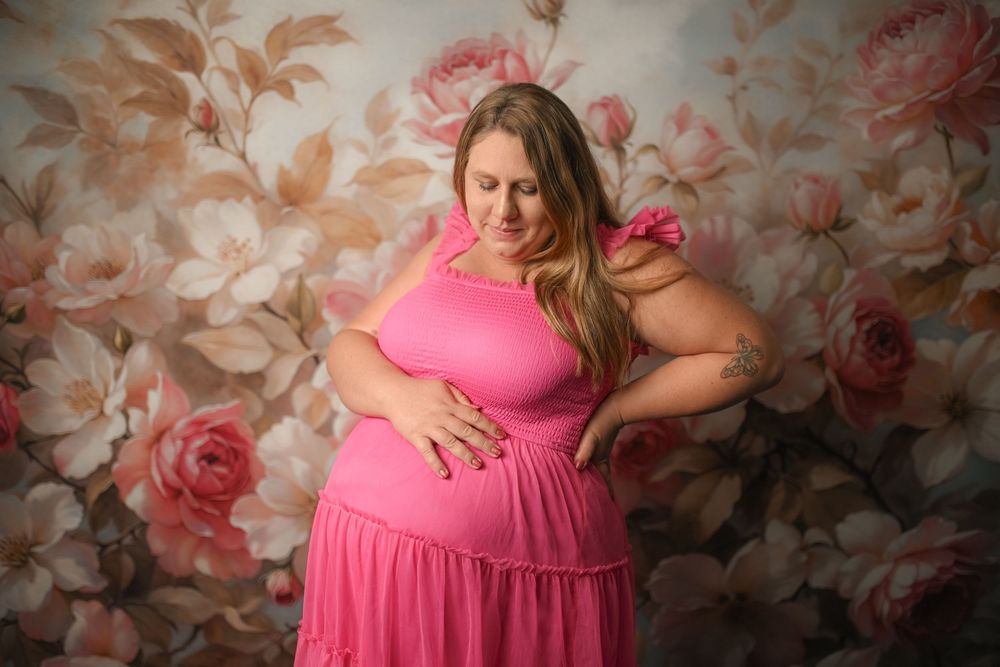 Pregnant woman in pink dress posing against floral backdrop during maternity photo session.